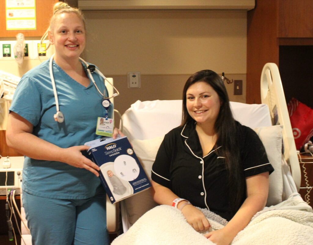 A nurse in blue scrubs stands beside a smiling patient in black pajamas sitting up in a hospital bed. The nurse is holding a box labeled “Snuggle Shield,” helping promote safe sleep while the patient rests under a white blanket.