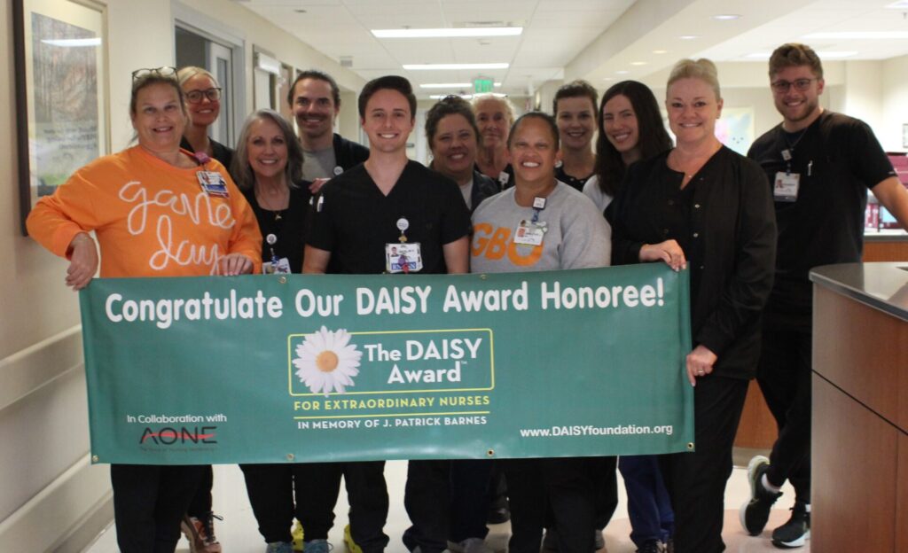 A group of smiling hospital staff stands in a hallway holding a green DAISY Award banner that reads, “Congratulate Our DAISY Award Honoree! The DAISY Award for Extraordinary Nurses.”.