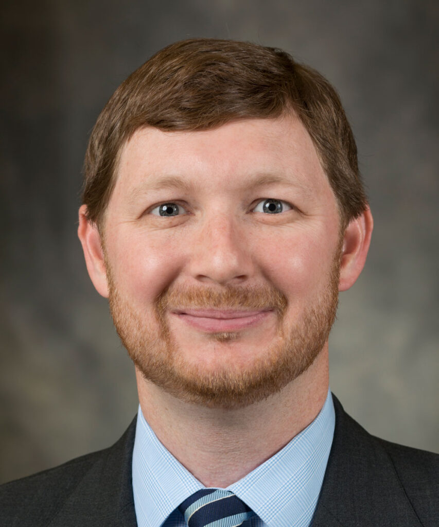 A man with short light brown hair and a trimmed beard, wearing a dark suit, light blue shirt, and striped tie, smiles at the camera against a blurred gray background—projecting confidence before his upcoming shoulder surgery.