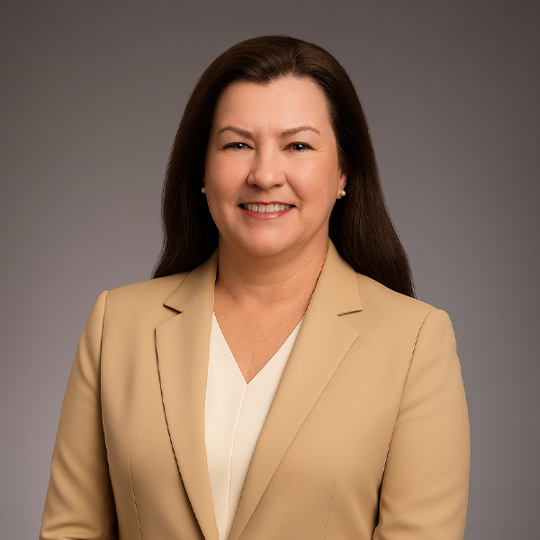 A woman with long brown hair, wearing a beige blazer over a white blouse, smiles at the camera against a plain, gray background.