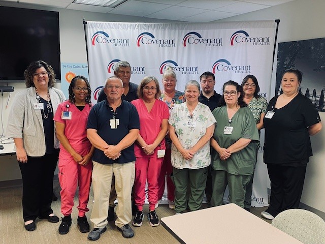 A group of twelve healthcare workers, some in scrubs and others in business attire, stand together smiling in front of a Covenant Health banner in a meeting room, gathering for their weekly updates.