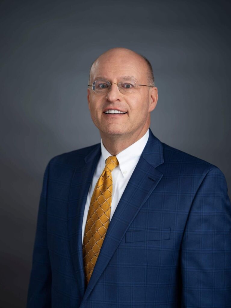 A man in glasses, a blue plaid suit, white shirt, and yellow patterned tie poses for a professional portrait against a plain dark background.