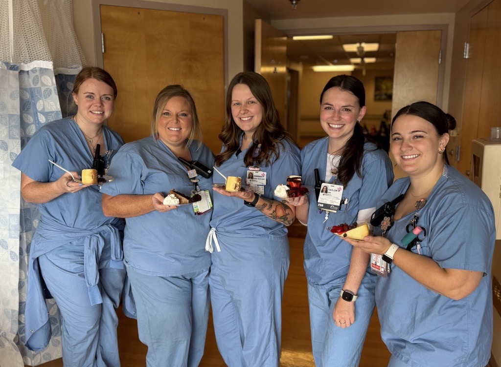 Five smiling women in blue scrubs stand together indoors, each holding slices of cake and wearing ID badges.