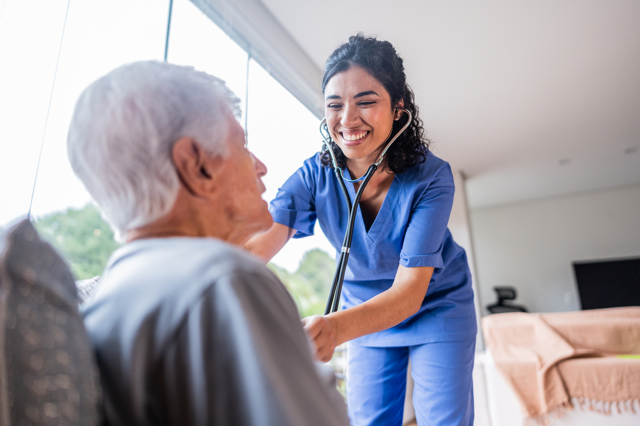 A smiling healthcare worker in blue scrubs uses a stethoscope to check the heartbeat of an elderly man.