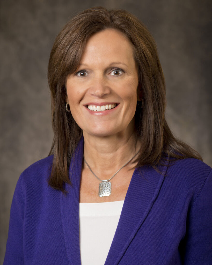 A woman with straight brown hair, wearing a purple blazer, white top, silver necklace, and hoop earrings, smiles at the camera in front of a blurred dark background, ready to share weekly updates.