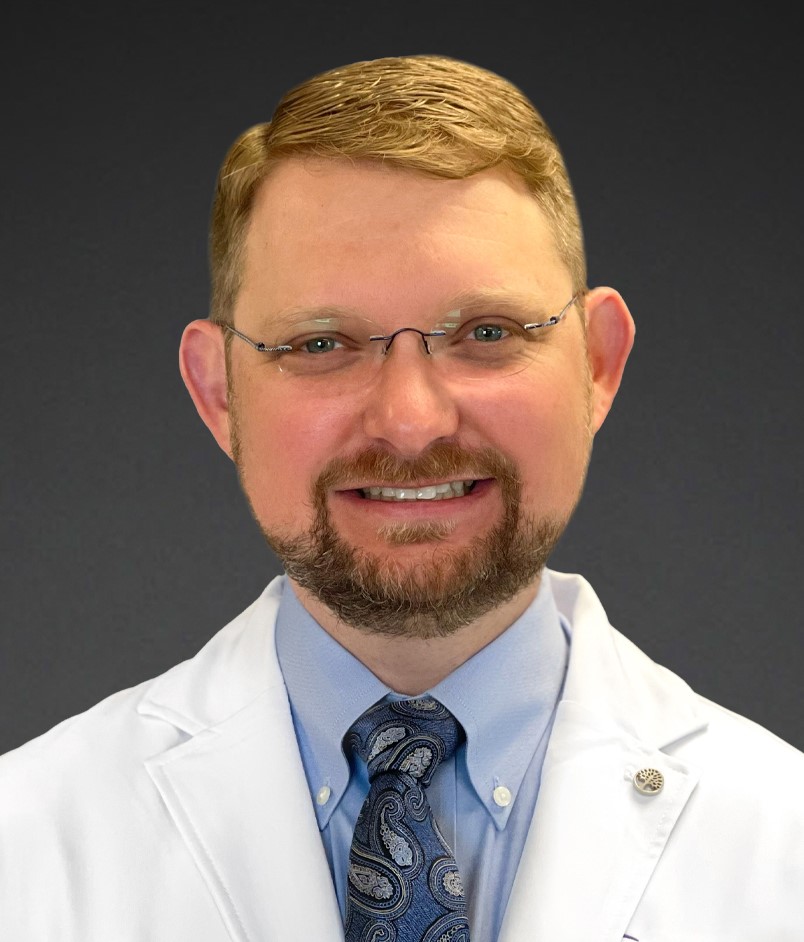 A man with light brown hair, glasses, and a beard, wearing a white medical coat, light blue shirt, and paisley tie, smiles confidently against a plain dark background&mdash;representing Advanced Care at Home.