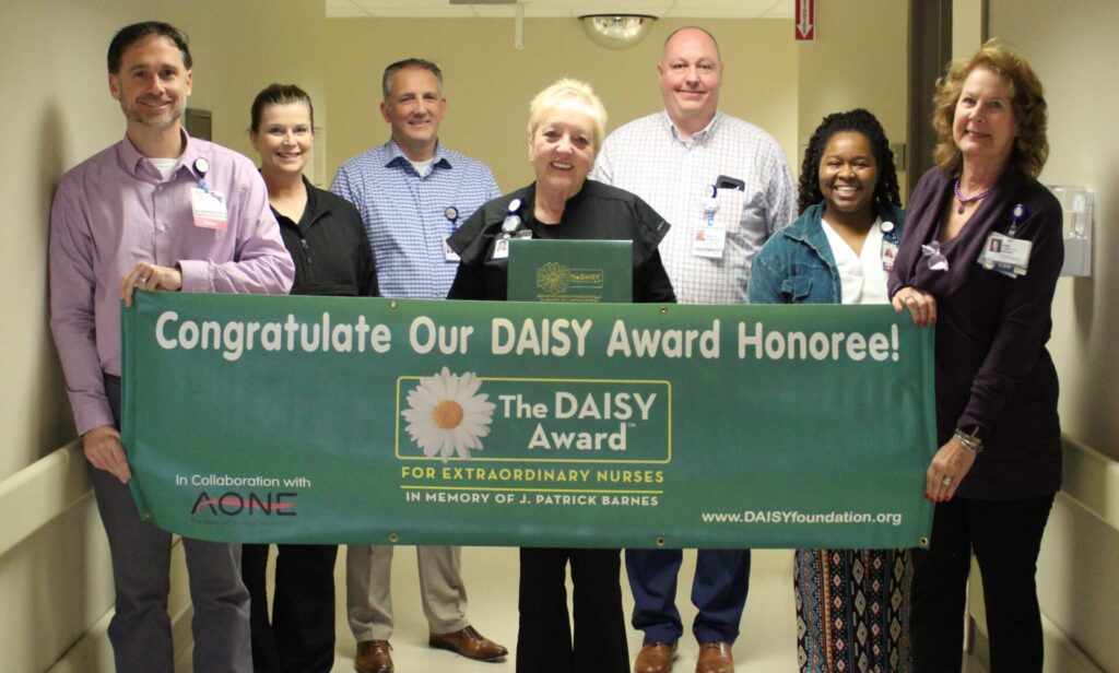 Seven smiling healthcare professionals stand in a hallway holding a large green banner that proudly reads, “Congratulations Our DAISY Award Honoree! The DAISY Award for Extraordinary Nurses.”.