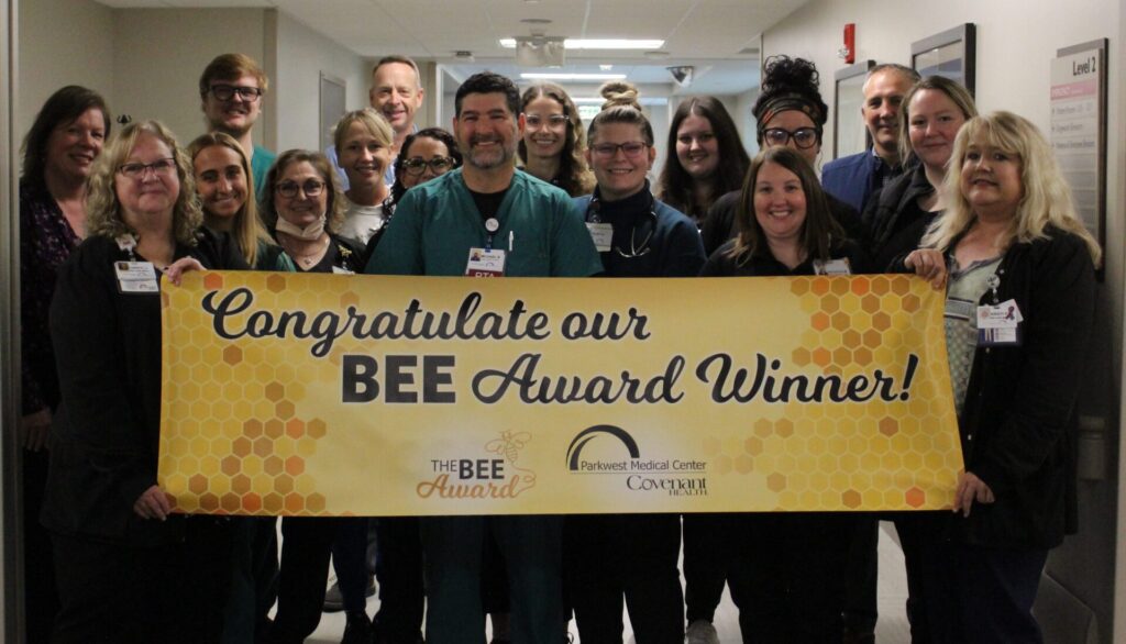 A group of smiling hospital staff stands in a hallway, proudly holding a yellow banner with honeycomb designs and hospital logos that reads, "Congratulate our BEE Award Winner!