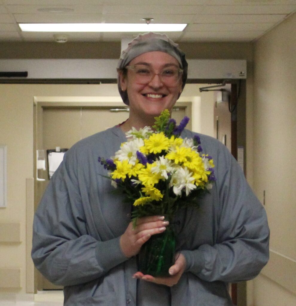 Monica in scrubs, wearing surgical cap, holding vase of daisies.