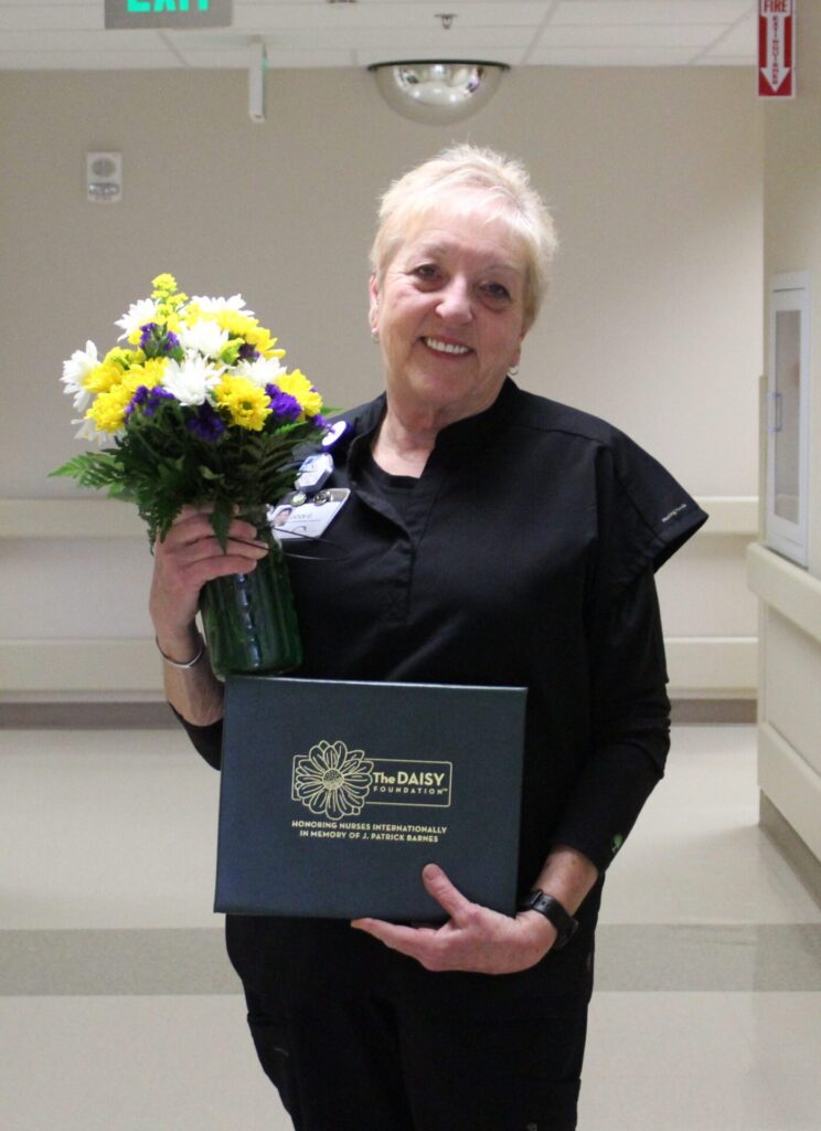 A woman in black scrubs smiles while holding a bouquet of flowers and a green folder featuring the DAISY Award logo, proudly standing in a hospital hallway.