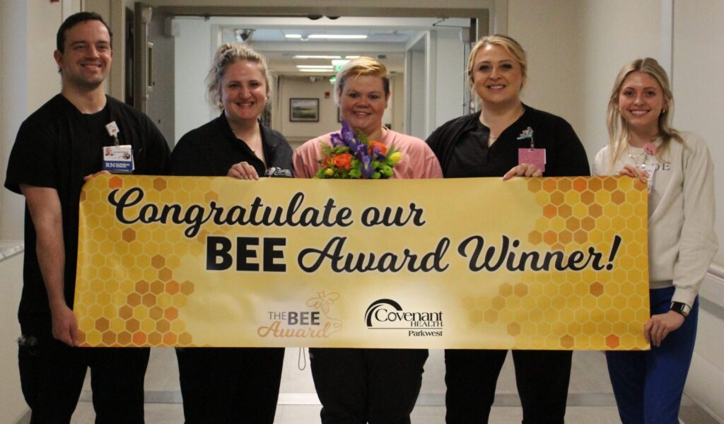 Five people stand in a healthcare facility hallway, smiling and holding a banner that reads "Congratulate our BEE Award Winner!" The proud BEE Award recipient at the center holds a bouquet of flowers.