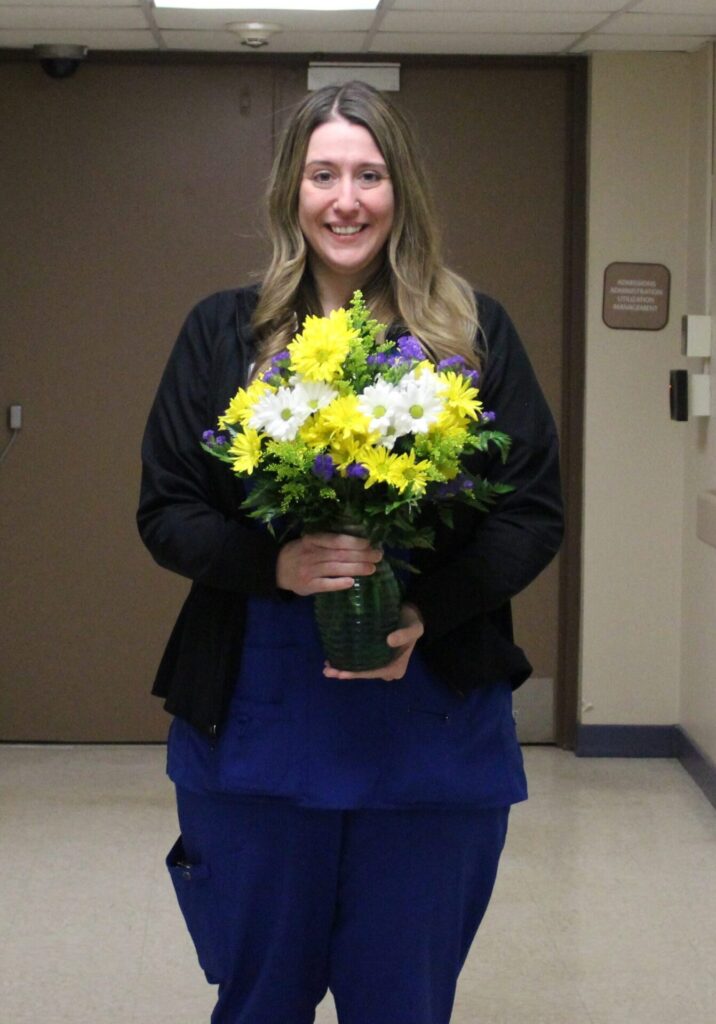 A woman with long, wavy hair smiles while holding a bouquet of yellow, white, and purple flowers in a green vase. Wearing blue scrubs and a black jacket, she stands in a hallway celebrating her DAISY Award honor.