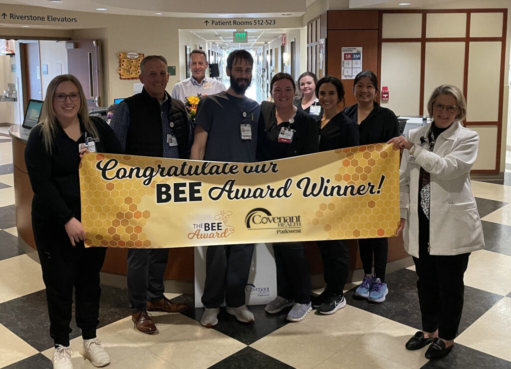 A group of hospital staff stand together in a hallway, proudly holding a large yellow banner that reads, Congratulations our BEE Award Winner! Covenant Health Partners. They are smiling and celebrating the BEE Award in this photo.