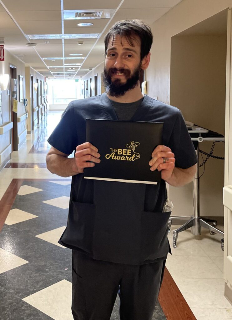 A man in dark scrubs stands in a hallway, smiling as he proudly holds his BEE Award certificate folder. Medical equipment can be seen in the background.