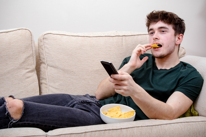young adult man eating chips on a couch watching tv