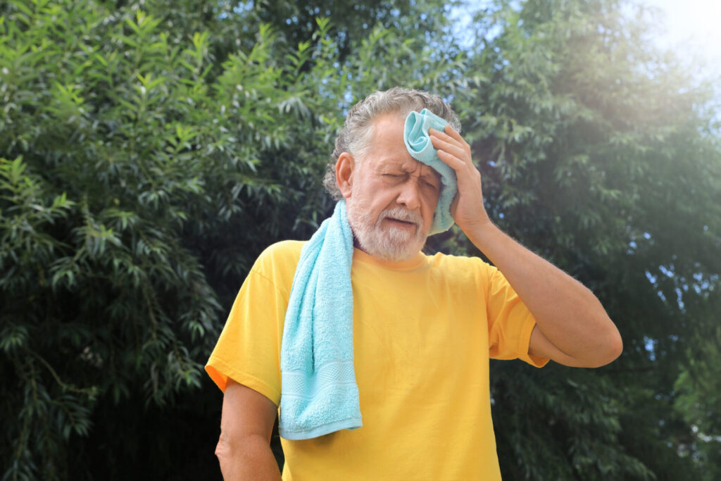 An older man in a yellow shirt stands outdoors, wiping sweat from his forehead with a towel. He looks tired and hot, with trees and sunlight in the background.