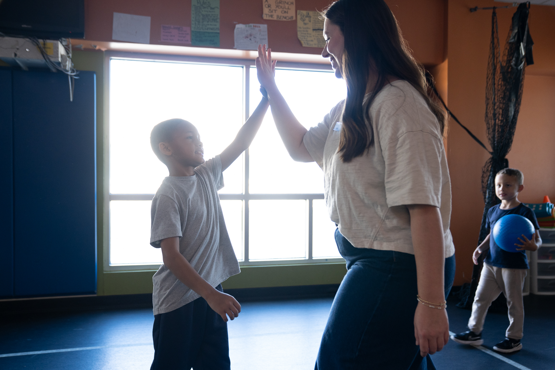 A woman and a young boy are giving each other a high-five in a brightly lit room, highlighting moments of youth development. Another child holding a blue ball stands in the background.