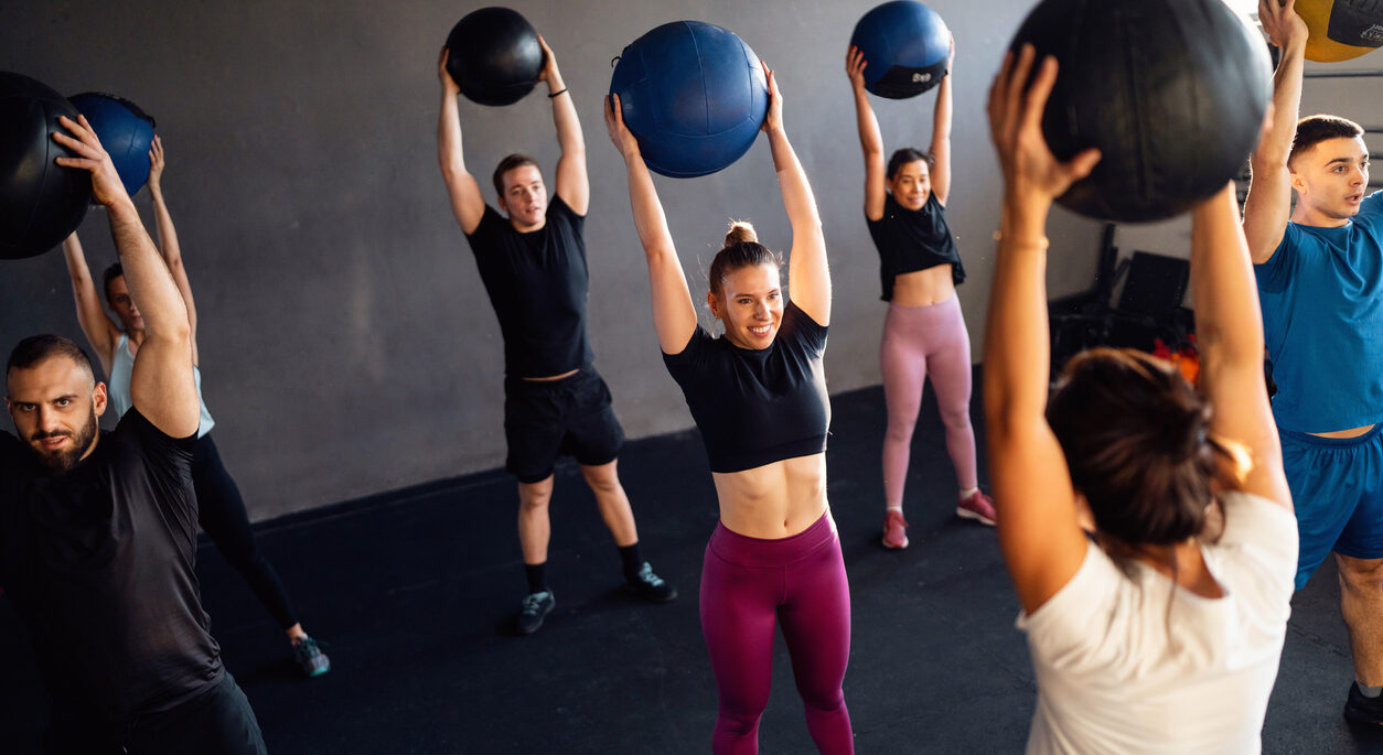 A group of young people with female cross trainer exercise with medicine weight balls during a group workout at the gym.