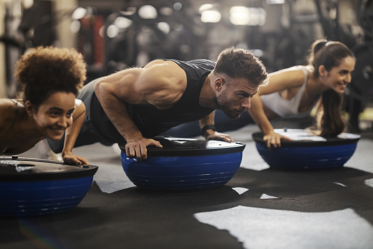 A fitness group of three people do pushups with bosu ball in a gym.
