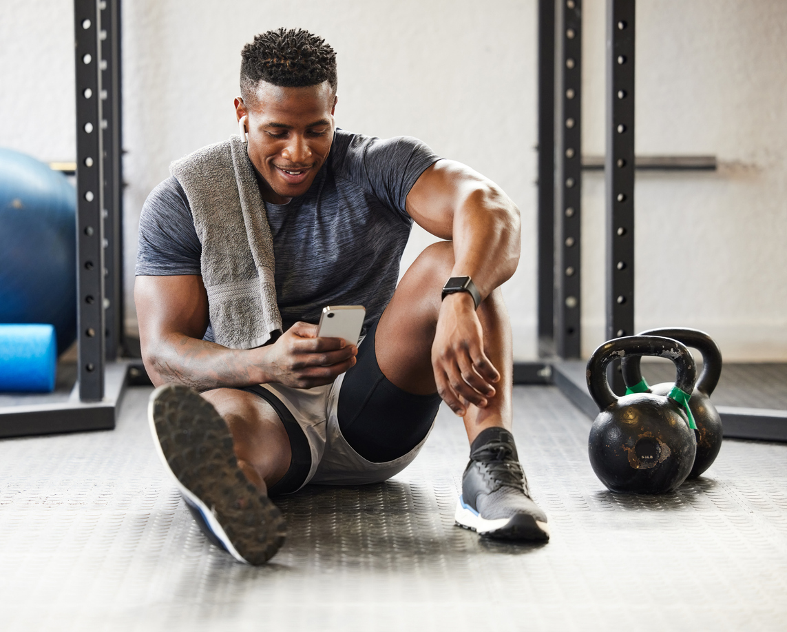 A muscular young mans sits and uses a cellphone between exercises at the gym.