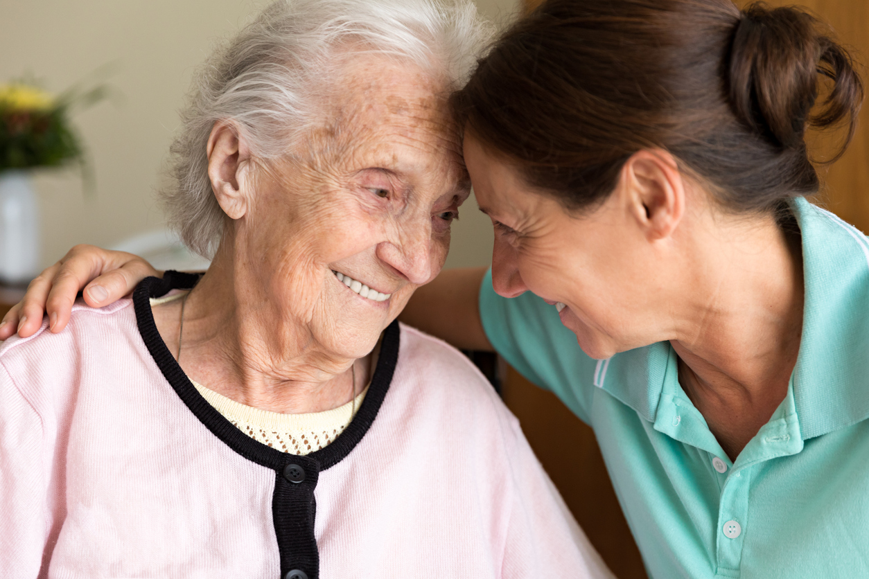 An elderly woman and a younger woman smile warmly, touching foreheads and embracing affectionately, conveying a sense of love and connection as they care for someone with dementia.