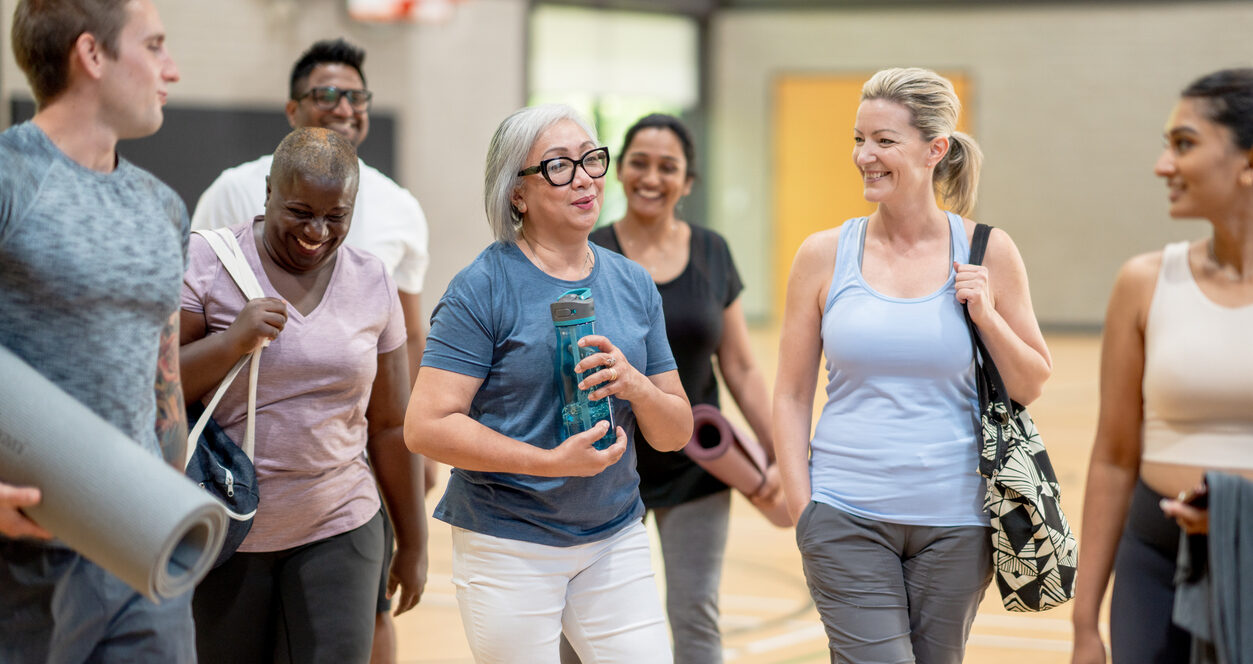 A diverse group of adults chat and laugh while prepping for an indoor group yoga class.
