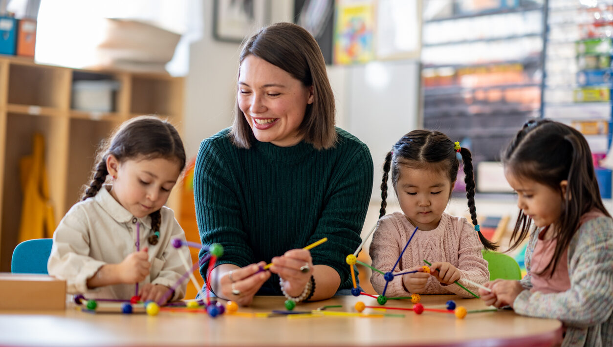 A teacher sits with a small group of young students at a desk as they play with various building materials.
