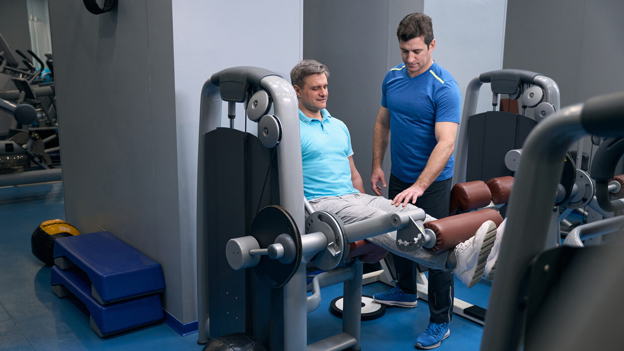 A fitness instructor assists a man with a seated leg extension on an exercise machine.