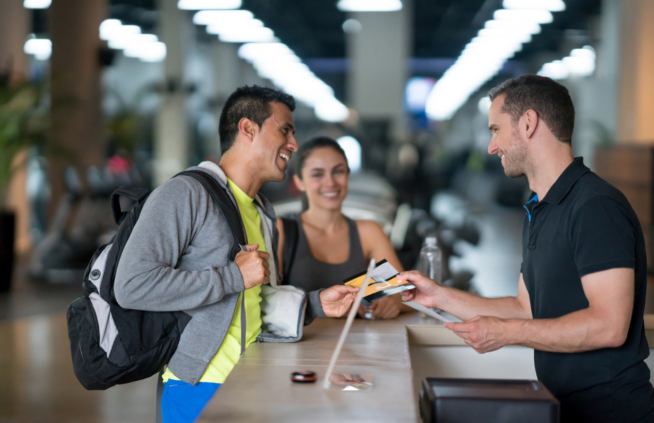 A smiling male receptionist checks two members into the fitness center.
