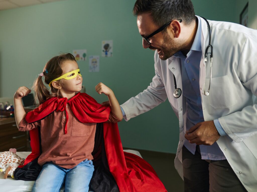 Female child wearing superhero cape and flexing muscles during visit to male physician