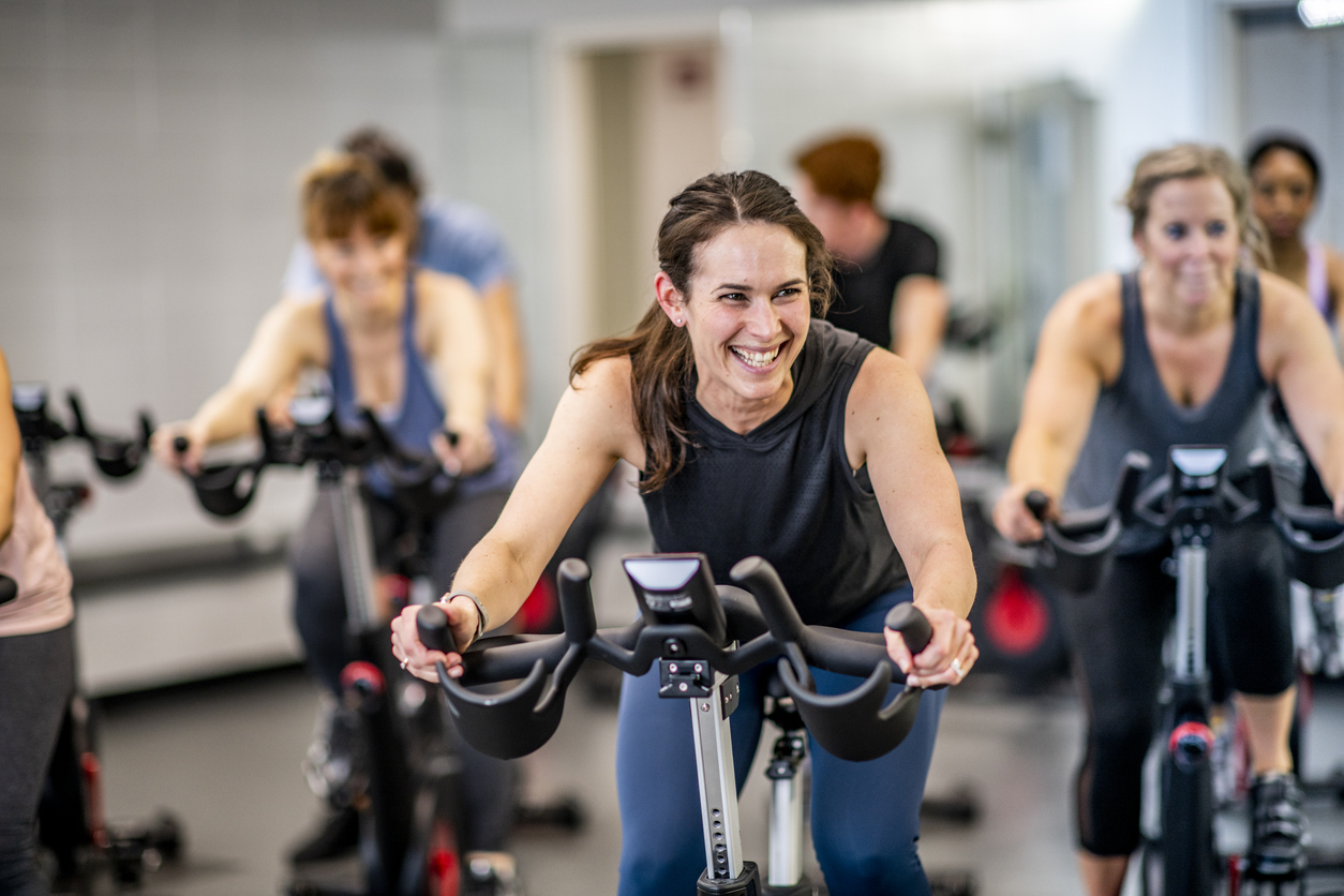 A woman smiles as she rides an exercise bike in an indoor cycling class at the gym.