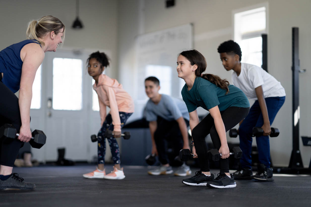 A young woman fitness instructor smiles as she teaches a group of kids an exercise with hand weights in a children's fitness class.