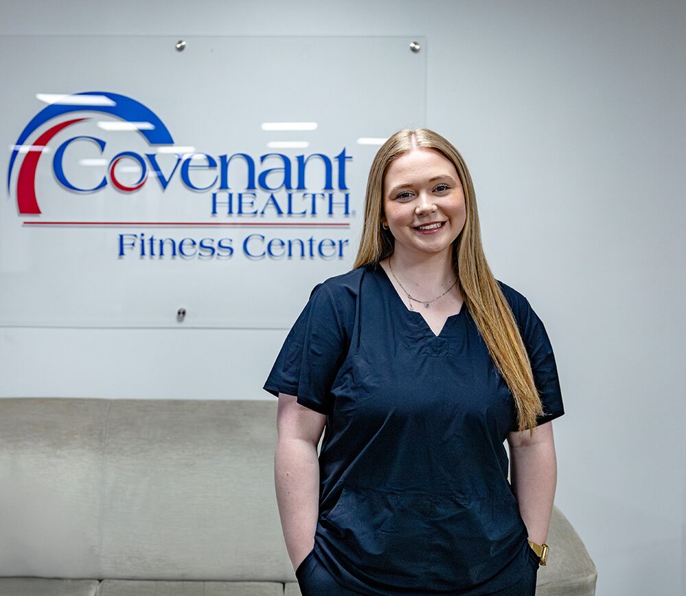 A woman in navy scrubs stands smiling in front of a “Covenant Health Fitness Center” sign inside a modern, clean office with a gray couch in the background.