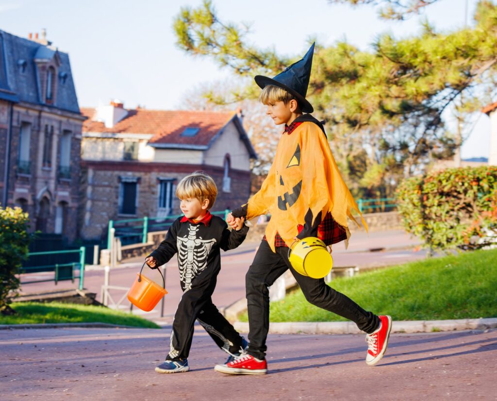 Young boy holding hand of older boy as they wear costumes and carry Halloween buckets and walk on a suburban street.