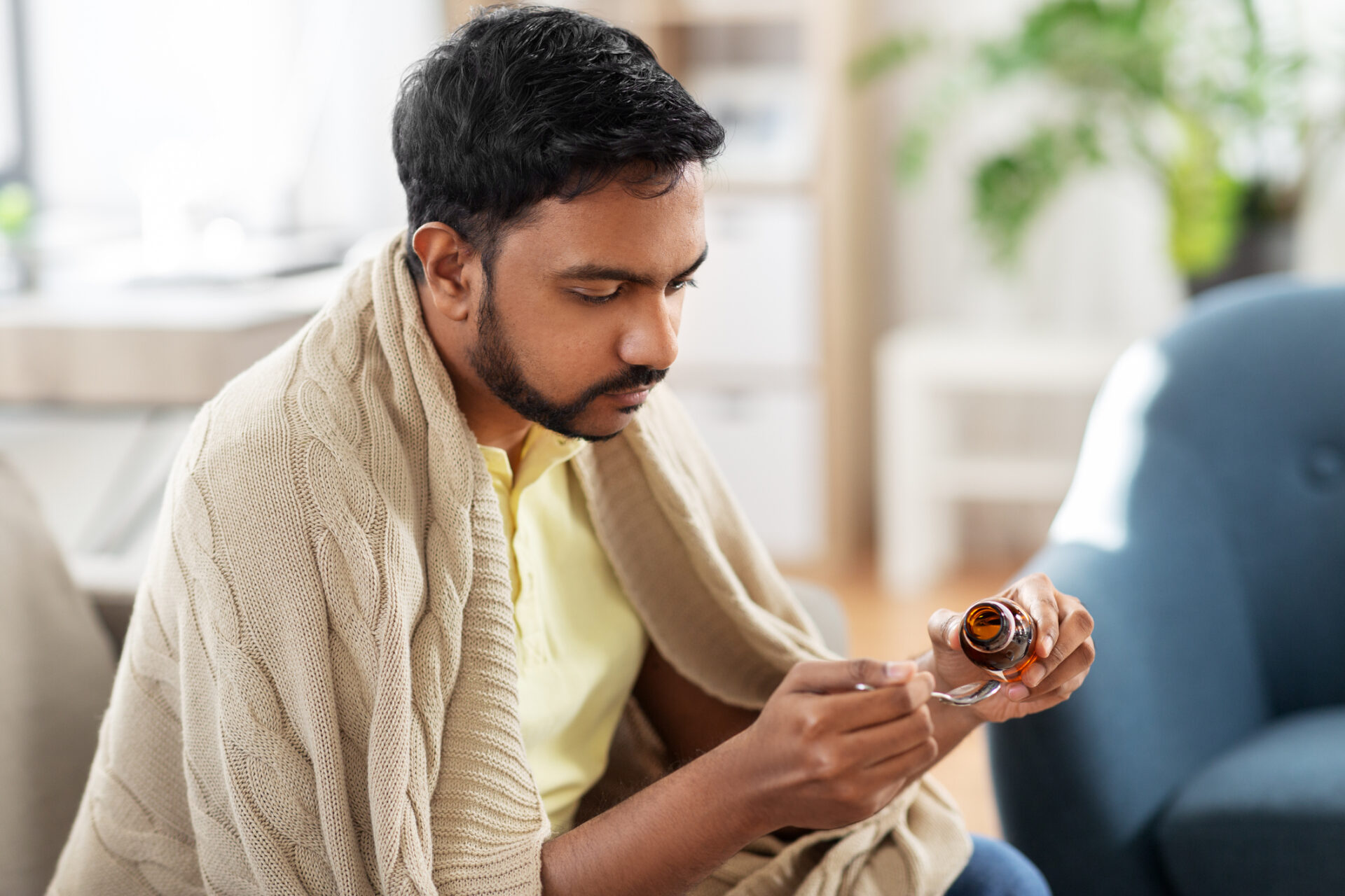 A man wrapped in a beige blanket sits on a couch, looking down as he pours the best cold medicine from a brown bottle onto a spoon, suggesting he is unwell and about to take his medication.