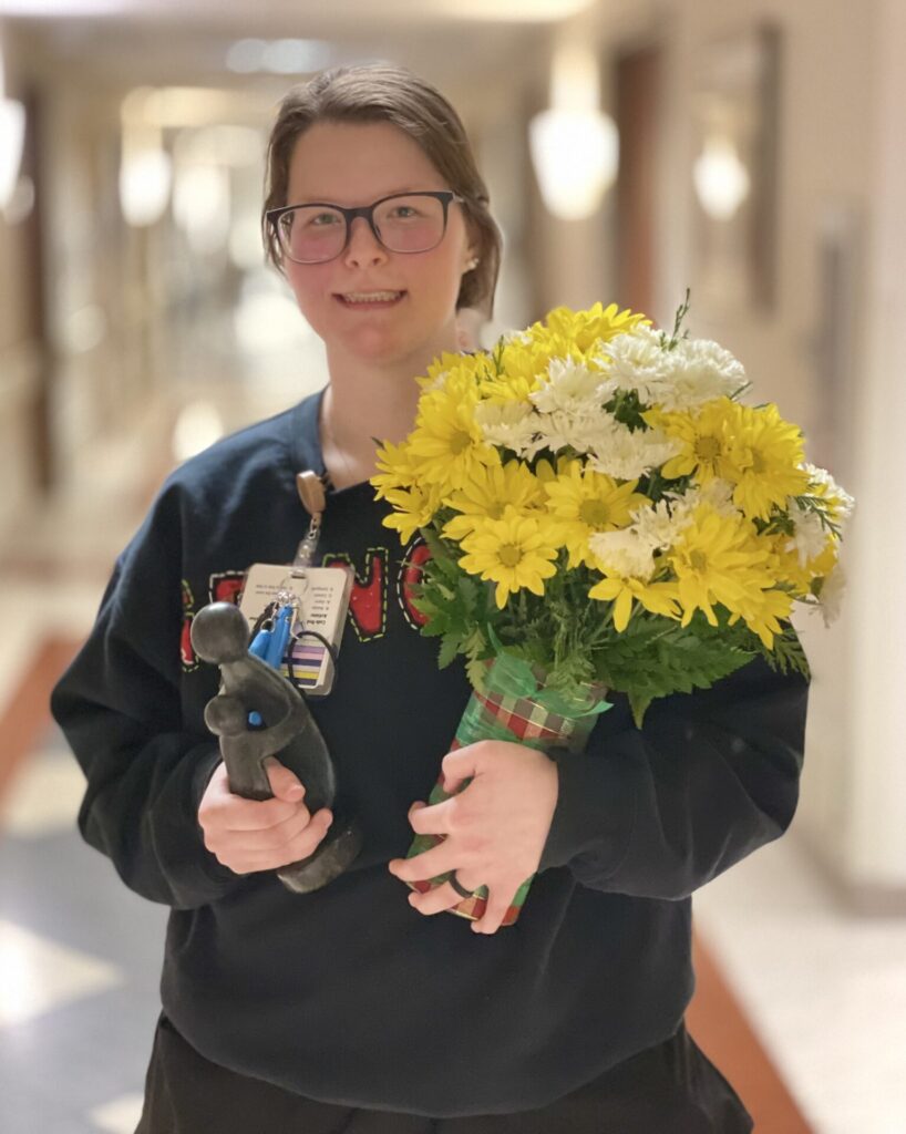 Sarah in black scrubs, smiling and holding daisies and sculpture.