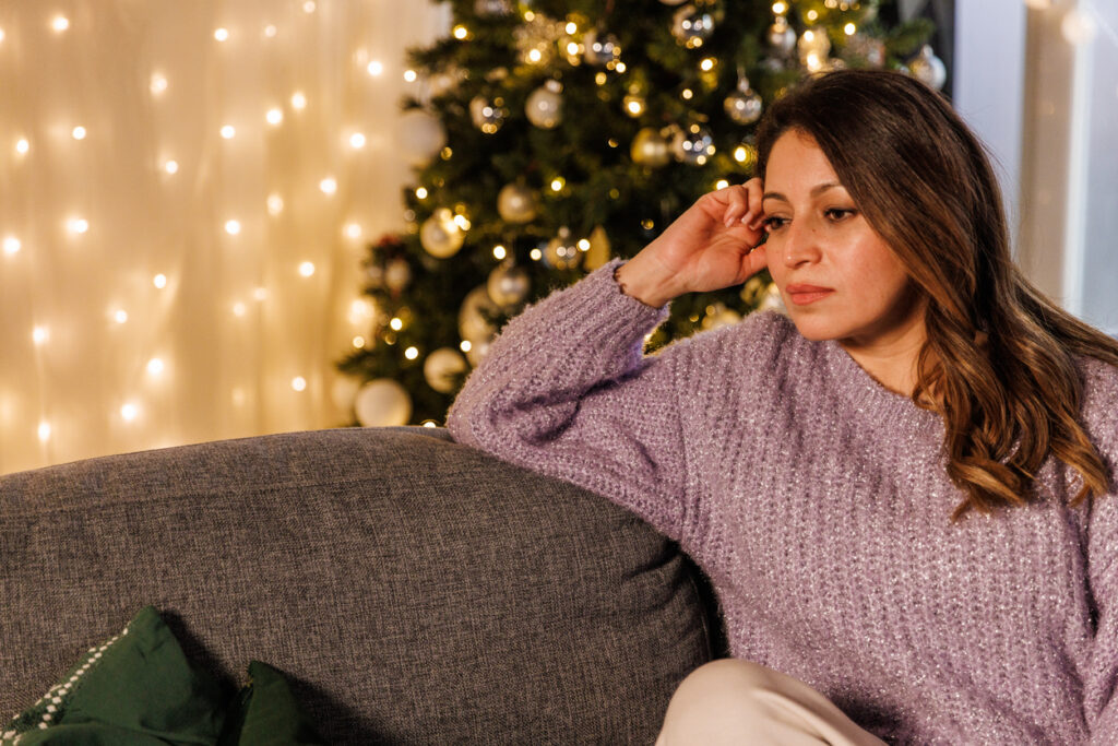 Woman looking pensive is seated on couch near Christmas tree. A gold curtain with sparkling lights is behind the couch and tree.