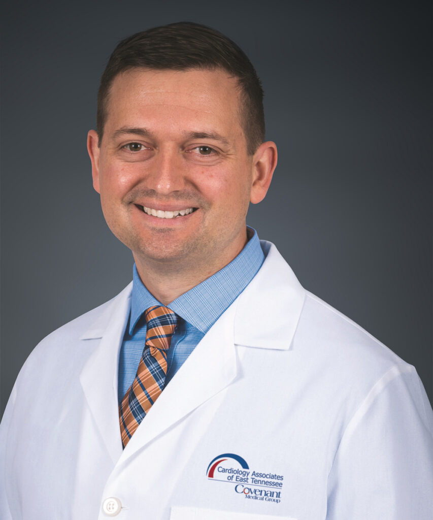 A smiling man wearing a white medical coat, blue shirt, and plaid tie, posing against a plain dark background. The coat features the logo for Cardiology Associates of East Tennessee.