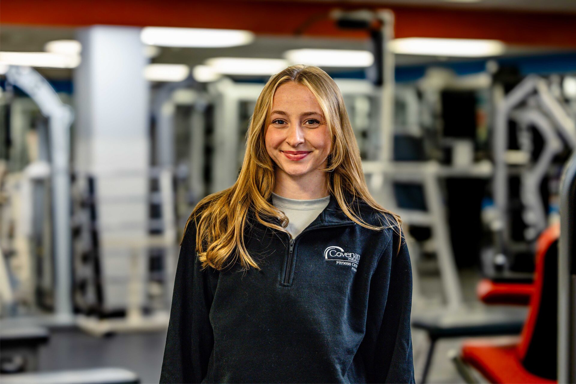 Untitled-1 A young woman with long blonde hair wearing a dark fleece pullover stands smiling in a gym filled with various exercise equipment. The background is softly blurred.