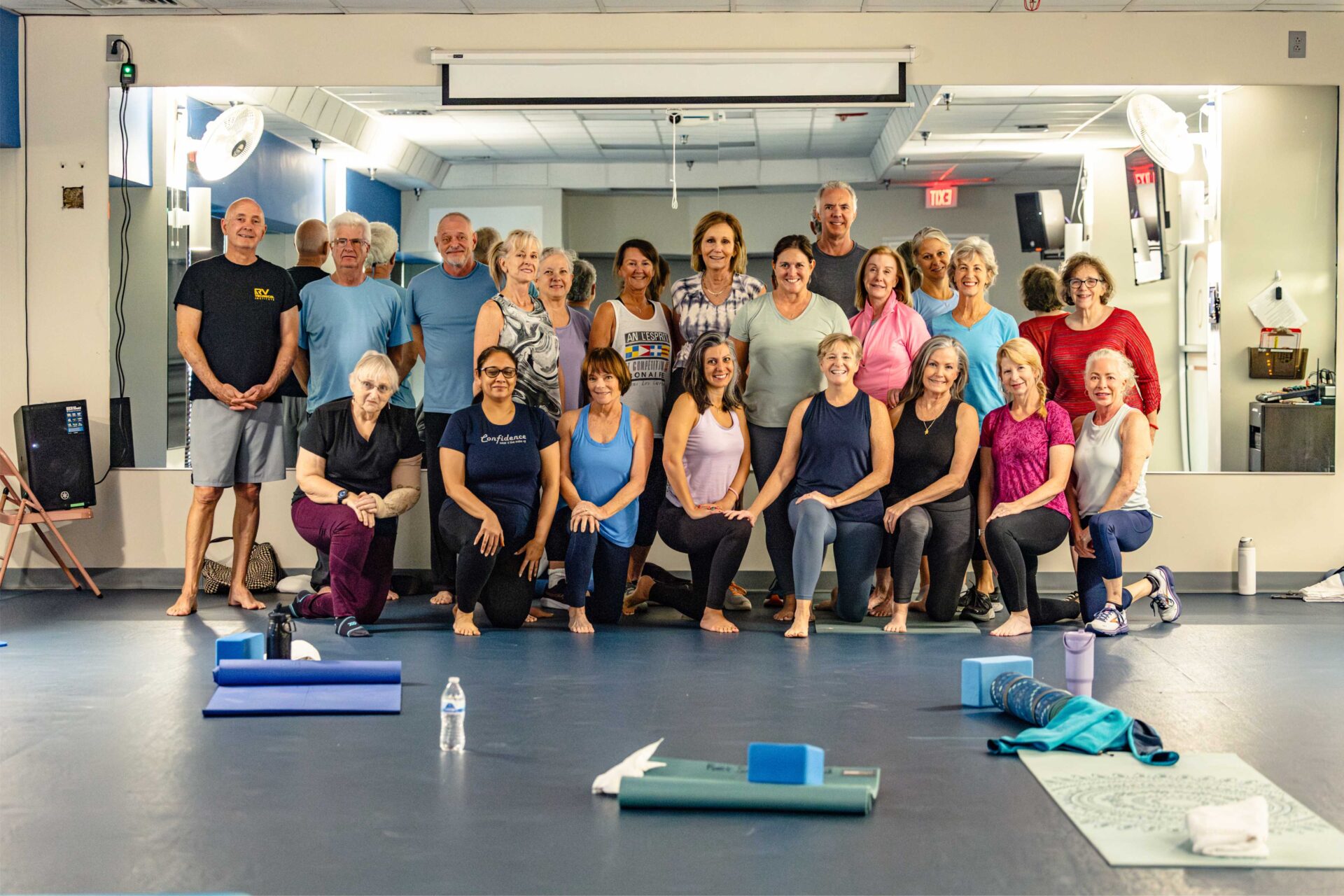 Untitled-4 A group of adults, men and women, pose and smile together in a yoga studio. Yoga mats and props are scattered on the floor, and a large mirror reflecting the group is on the back wall.