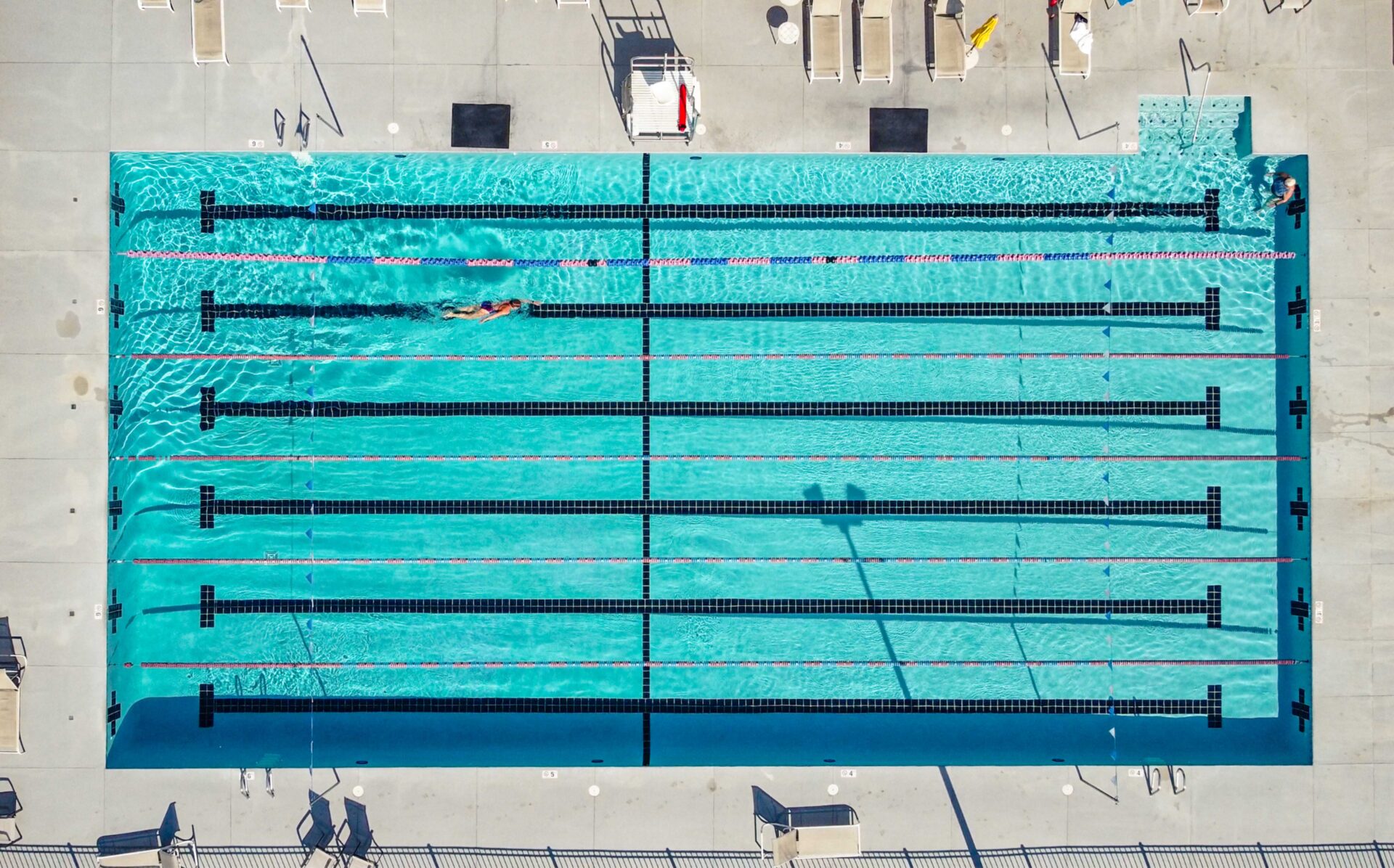 Untitled-81 Aerial view of an outdoor swimming pool with six lanes, one person swimming in a lane, empty poolside chairs, and clear blue water reflecting sunlight.