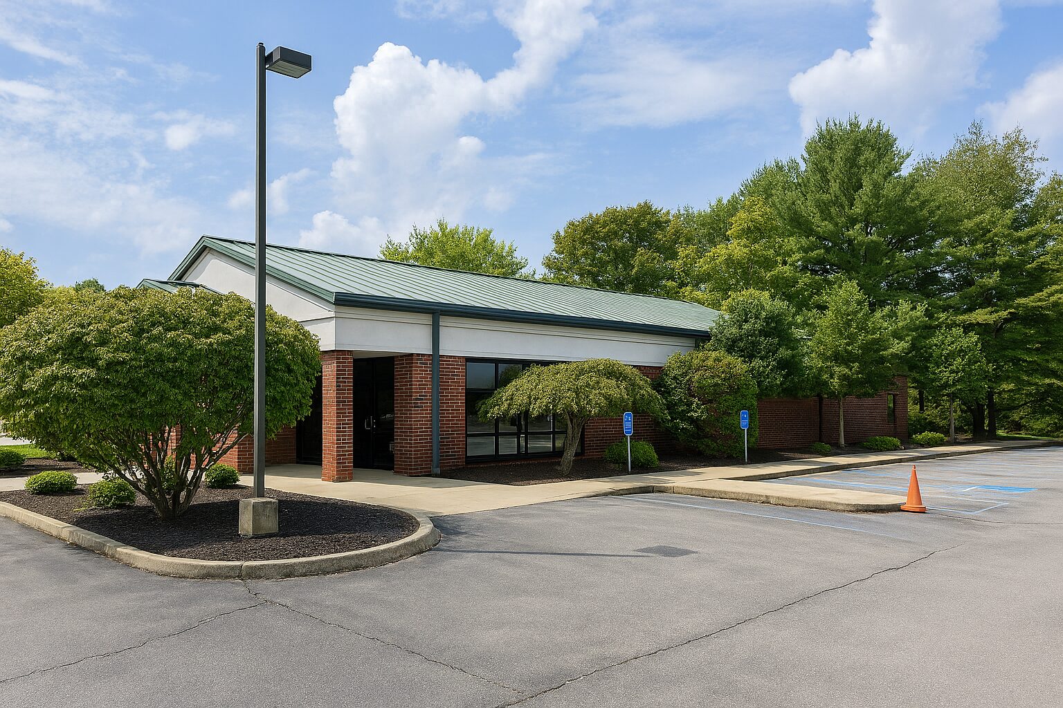 A single-story brick building with a green roof, surrounded by trees and bushes. There is a mostly empty parking lot with a few handicapped spaces and an orange traffic cone near the entrance.