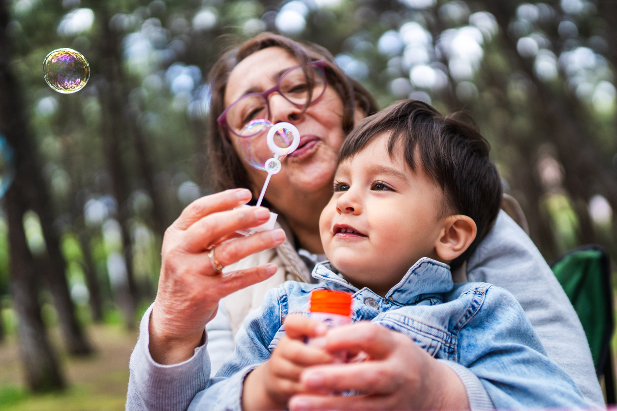 An older woman and a young child sit outdoors among trees, blowing soap bubbles together—a joyful reminder of the simple pleasures celebrated through quality pulmonology services. The woman holds the bubble wand while the smiling child watches a bubble float nearby.