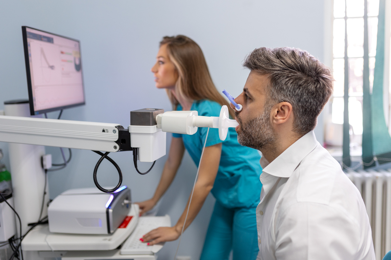 A man undergoes a lung function test, breathing into a spirometer, while a medical professional reviews data on a computer in a bright clinic room offering advanced pulmonology services.