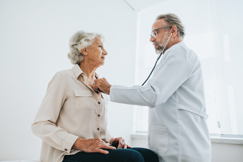 Physician is standing next to a seated older woman and is using a stethoscope to listen to her heart