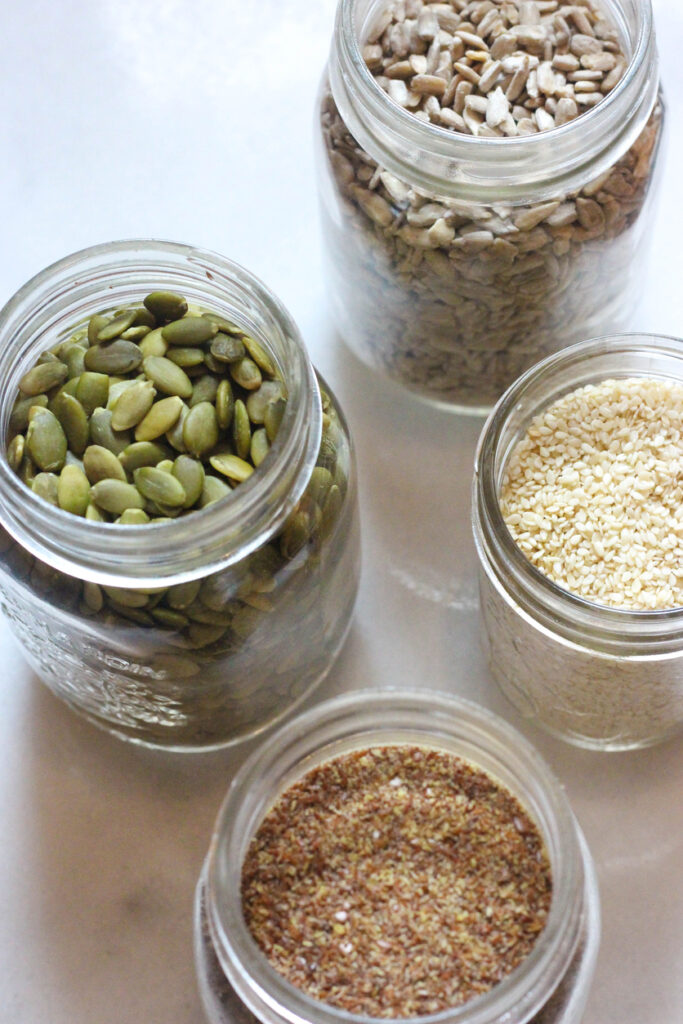 Photo of four jars of different seeds. Seed cycling is a trending nutrition approach to help with luteal phase symptoms.