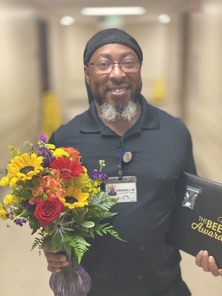African American man with glasses, smiling, holding a bouquet of flowers and an award.