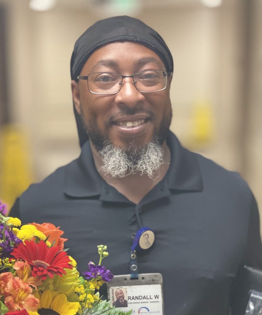 African American man with glasses, smiling, holding a bouquet of flowers and an award.
