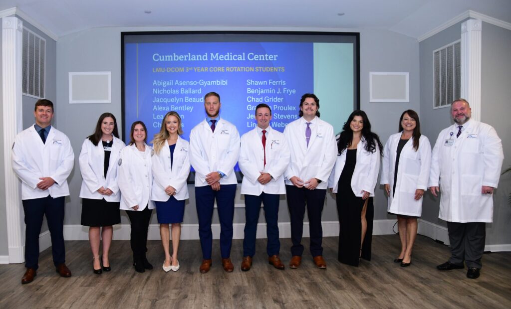 Five women and five men standing and smiling in front of screen titled Cumberland Medical Center LMU-DCOM Core Rotation Students. All are wearing white medical coats.
