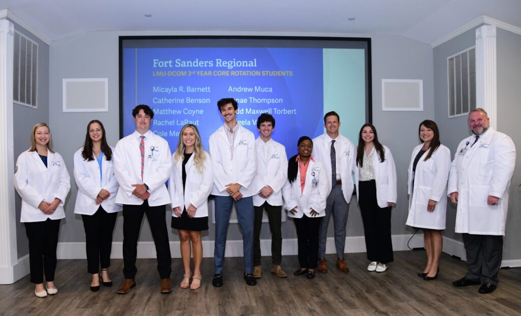 Six women and five men standing and smiling in front of a screen titled Fort Sanders Regional. All are wearing white coats.